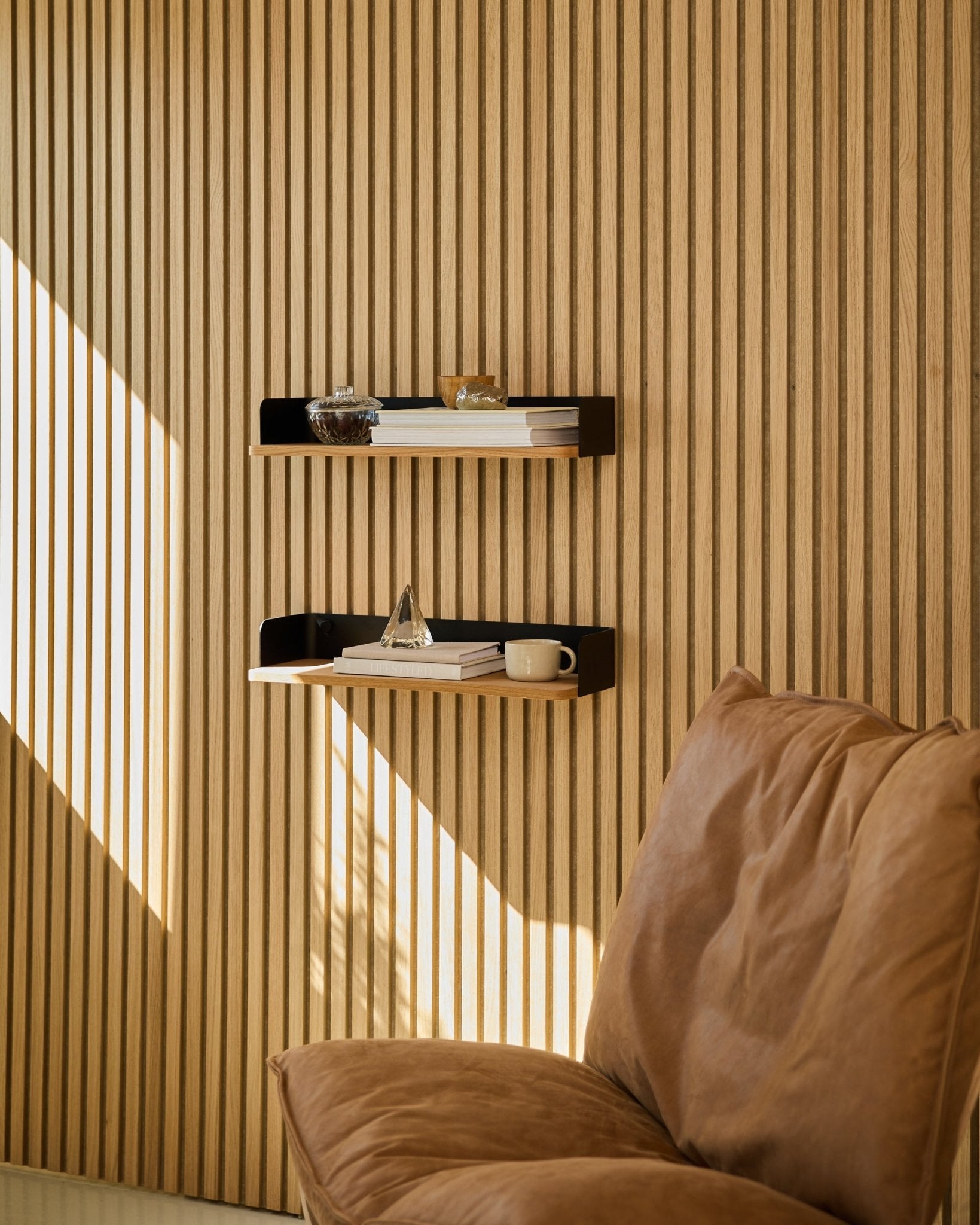 Brown Stuhl neben dem WoodUpp Linn Bücherregal in Natural Oak, mit Büchern und Deko im natürlichen Licht vor einer nordischen Holzlamellenwand Akustikpaneel.